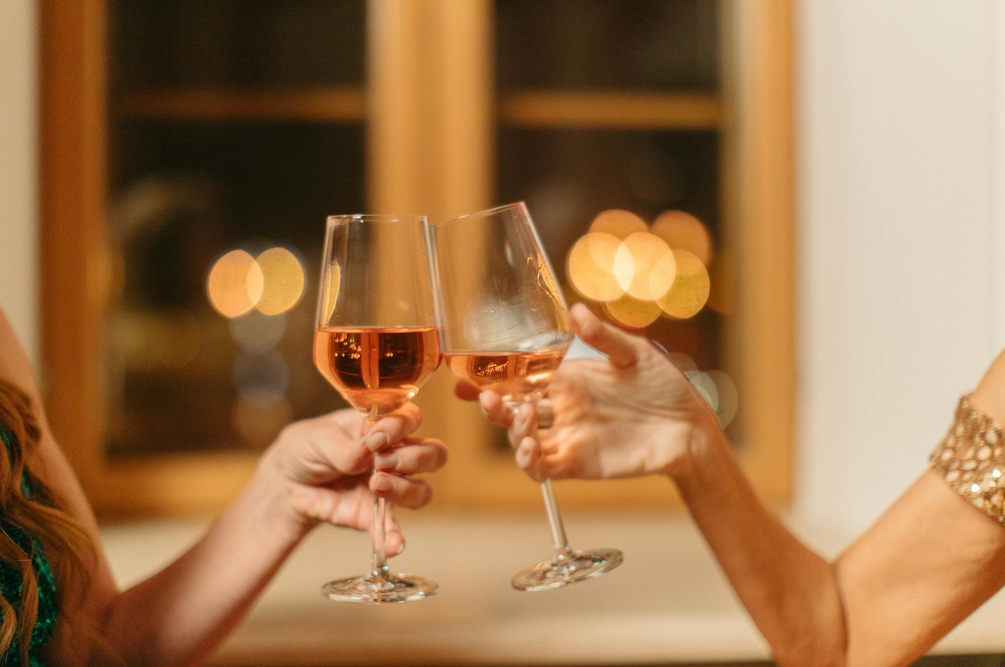 Close-up of two people toasting with rose wine glasses in a warm indoor setting.