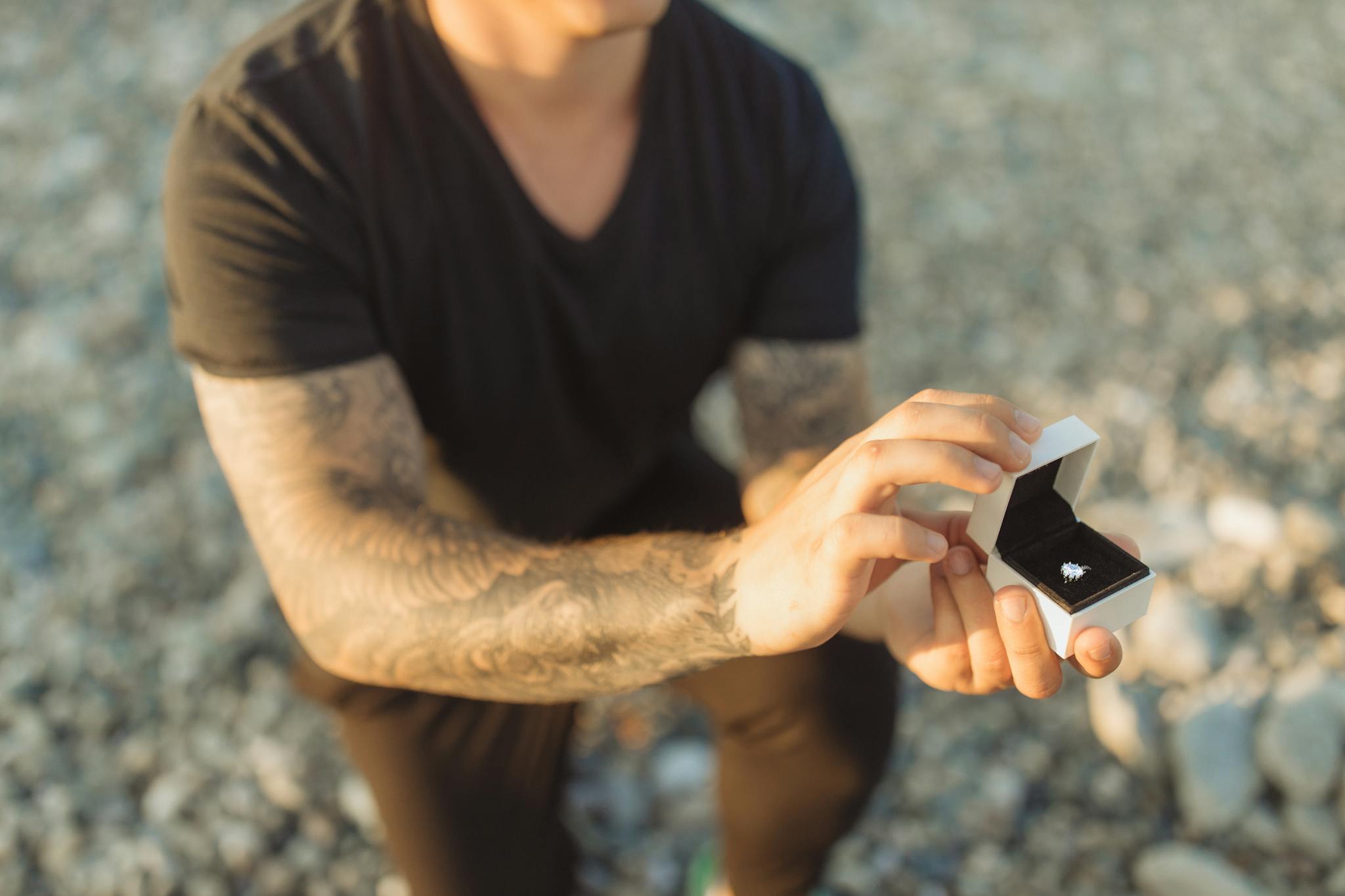 Man kneeling on a beach, presenting a diamond ring in a proposal scene.